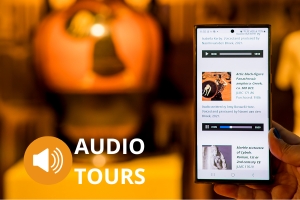 A phone showing a list of online audio descriptions with objects in the background. A speaker symbol in the bottom corner is next to text reading "Audio Tours."