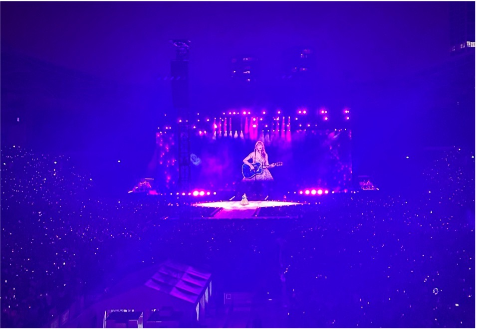 Taylor Swift preforms on stage in a stadium holding a guitar.