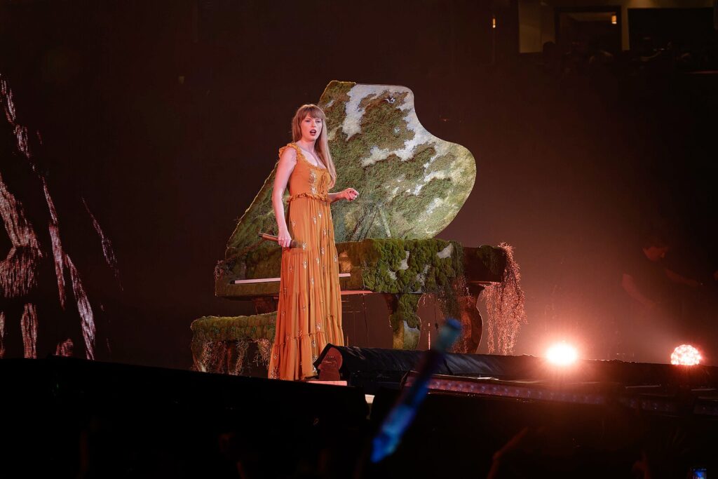 Taylor Swift stands on stage next to a grand piano looking out towards the audience.