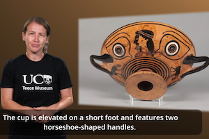 Jenna Holland wears a black Teece Museum t-shirt and stands in front of an image of an eye cup, ready to use NZSL to introduce the artefact.