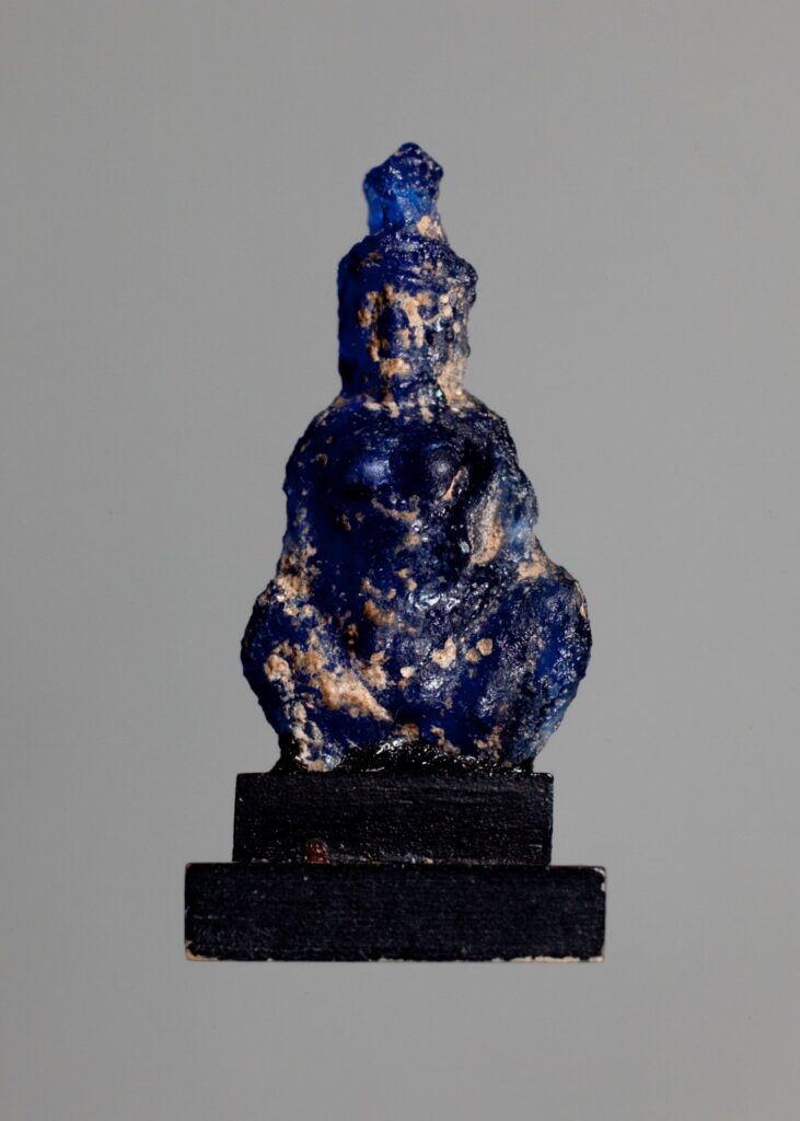Small blue glass amulet showing a woman squatting with her hands on her belly. The amulet is mounted on a black wooden base.