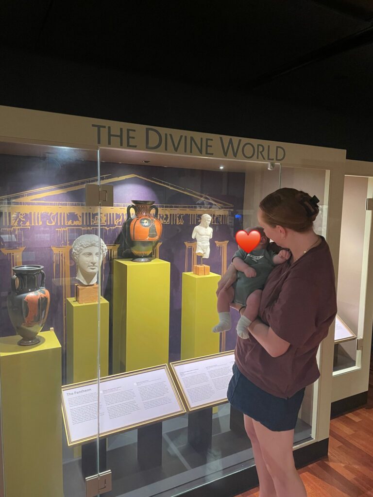 Emily Rosevear stands in the Teece Museum holding her baby. They are looking at a case of antiquities.