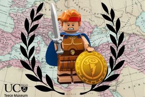 Image depicts a LEGO Herakles holding a sword and shield with a fierce look on his face. He is set against a map backdrop and encircled by a black wreath. the UC Teece Museum logo is in the bottom left corner.